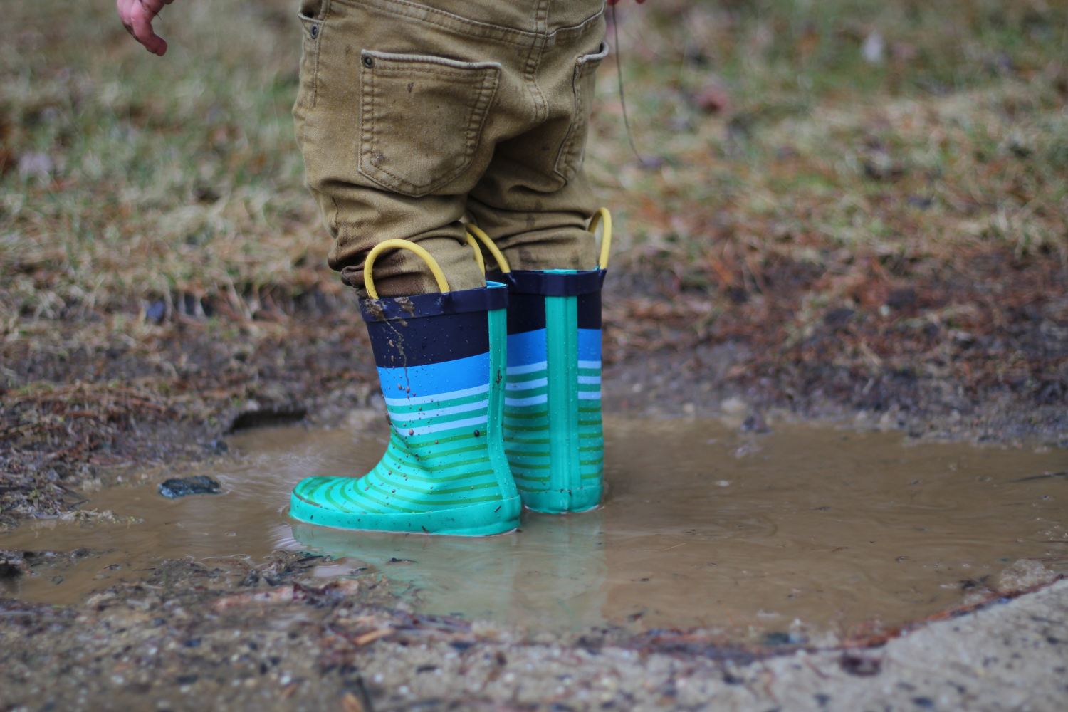 Kid with small green rubber boots playing in mud puddle.